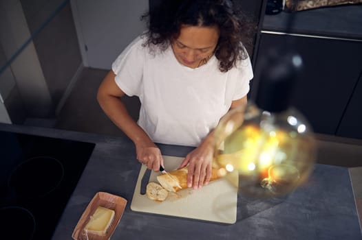 Top view through stylish vintage lamp on happy young woman, housewife standing at kitchen counter and cutting bread on a cutting board, making sandwiches with cheese for snack at home kitchen interior