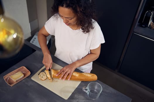 View from above of a woman using kitchen knife, cutting a loaf of whole grain French baguette bread on the cutting board, preparing delicious sandwiches with cheese for breakfast in the home kitchen.