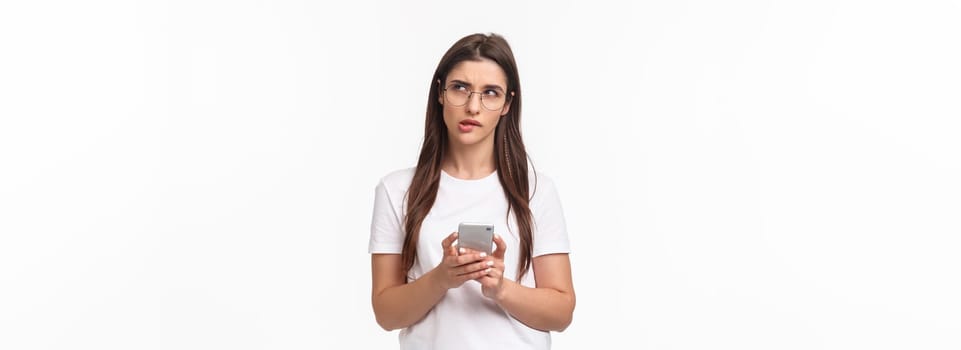 Communication, technology and lifestyle concept. Portrait of thoughtful young woman working remote, answer clients on phone, hold smartphone look up thinking what text back.