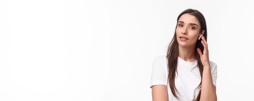 Close-up portrait of feminine attractive young woman in t-shirt, touching wireless earbud, listening music in headphone, looking camera, turn-on podcast or relaxing playlist, white background.