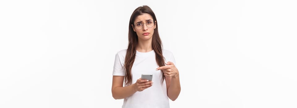 Communication, technology and lifestyle concept. Waist-up potrait of sad and gloomy young uneasy caucasian woman in glasses, frowning and pointing at mobile phone with regret or sadness.