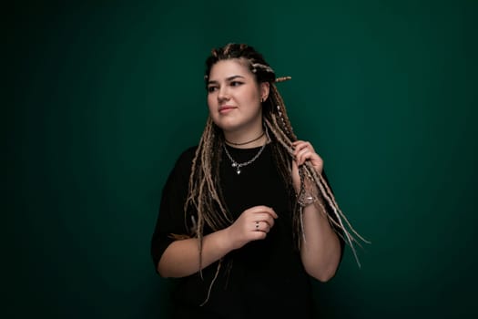 A woman with dreadlocks stands confidently in front of a solid green background. Her hair is styled in long, thick dreadlocks, and she wears a casual outfit. The setting is simple, focusing on the woman and her distinctive hairstyle.