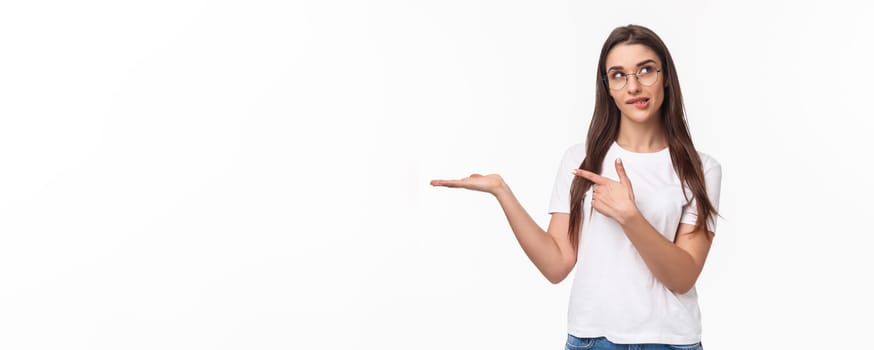 Waist-up portrait of thoughtful, pretty young woman holding arm as if have something lying in it, pointing at product with pondering face, bite lip look up thinking, white background.