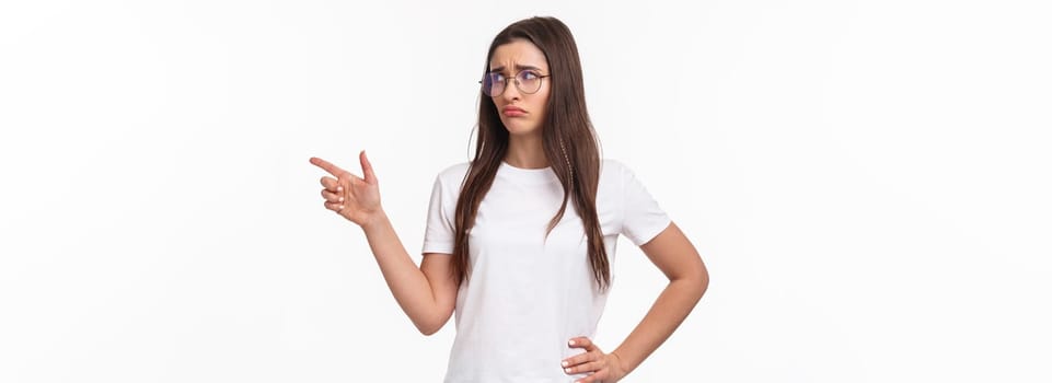 Waist-up portrait of uneasy, sad and gloomy young brunette woman in glasses, looking and pointing finger right with sulking unhappy face, express jealousy or regret, grieving white background.