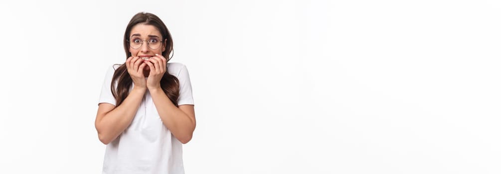 Waist-up portrait of scared young brunette girl in panic, exams are near feel frightened, biting fingers and staring terrified, trembling fear, standing white background afraid.