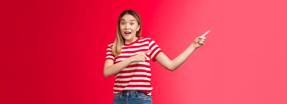 Hey hurry up look. Enthusiastic excited cute asian blond urban girl pointing left showing incredible offers, raise eyebrows thrilled talk camera smiling delighted stand red background impressed.