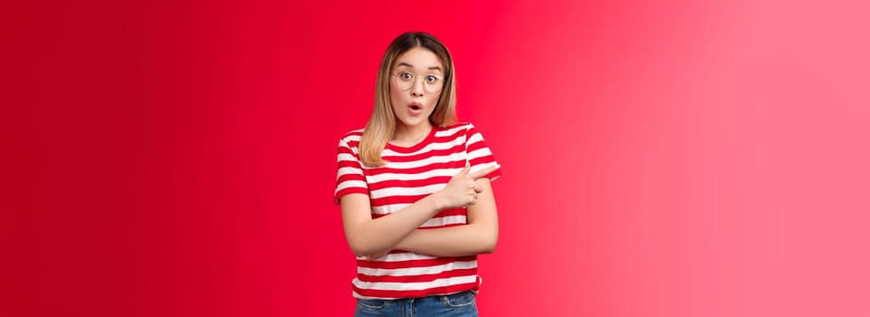 Fascinated surprised cute asian girl in glasses talking about incredible offer folding lips gasping, look wondered camera raise eyebrows astonished pointing left questioned intrigued, red background.