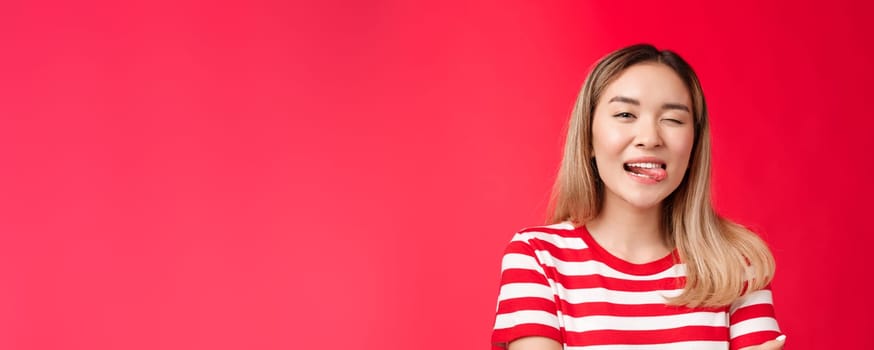 Close-up sassy carefree outgoing urban asian female student enjoy summer vacation wear striped t-shirt, show tongue playfully winking camera, stand red background positive friendly mood.