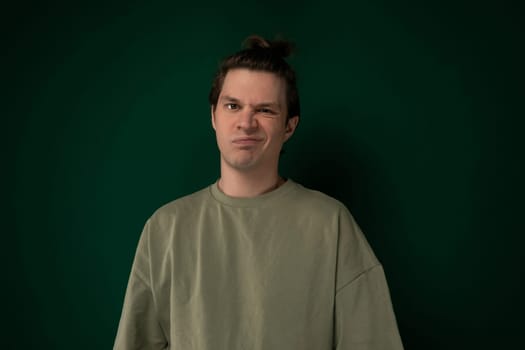 A man is standing in front of a bright green wall, looking straight ahead. He is wearing casual clothing and appears to be deep in thought, with his hands in his pockets. The wall provides a striking contrast to his dark clothing.