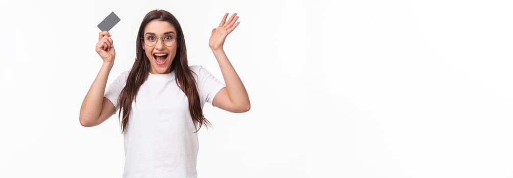 Waist-up of enthusiastic, happy surprised young woman reacting to wonderful news, raising hands up in amazement, holding credit card, receive salary, got her first paycheck, hurry to order something.