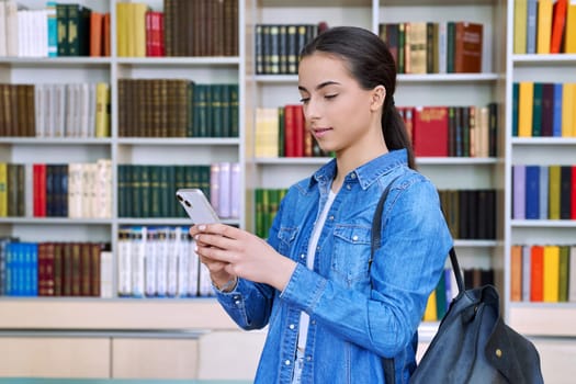 Teenage female student with backpack using smartphone, inside high school building, in library. Technologies, mobile educational apps applications, services, e-learning concept