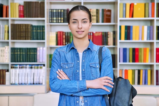 Portrait of smiling teenage girl student. Confident female teenager 16,17 years old with backpack, crossed arms inside high school building, background of library shelves books. Education, adolescence