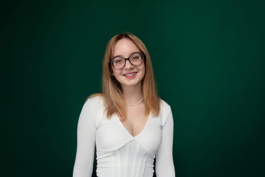 A woman with glasses is standing wearing a white dress. She looks directly at the camera, her hands by her sides. The background is plain and neutral.