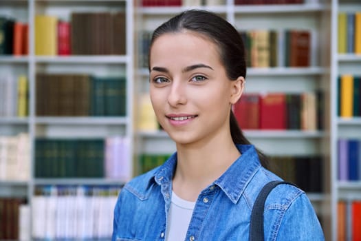 Headshot portrait of smiling teenage girl student. Confident female teenager 16,17 years old with backpack inside high school building, background of library shelves books. Education, adolescence