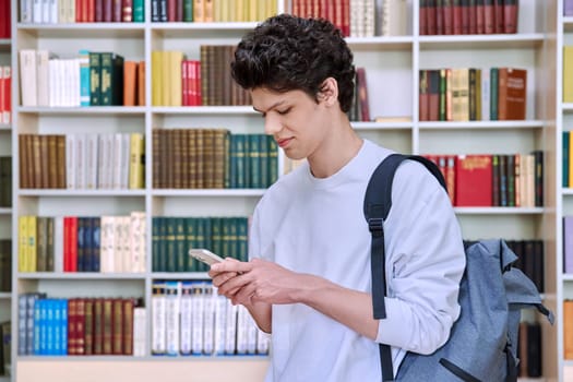 Teenage male student with backpack using smartphone, inside college building, in library. Technologies, mobile educational apps applications, services, e-learning concept