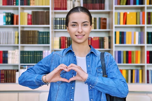 Happy teenage girl student showing heart gesture at camera with hands inside high school building. Joyful positive teenage girl 16, 17 years old in library. Emotions, education, lifestyle, adolescence