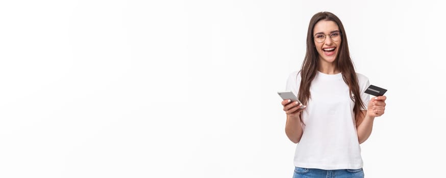 Portrait of happy smiling pretty woman in glasses holding credit card, mobile phone, laughing at camera easily paying for her purchase online store, buy things with application, white background.