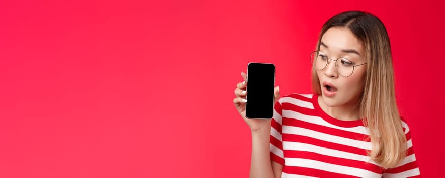 Close-up wondered surprised attractive asian blond girl wear glasses hold smartphone looking impressed amused phone screen, open mouth interesting app telephone display, stand red background.