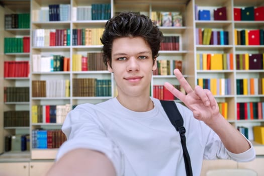 Selfie photo portrait of smiling handsome college student guy showing hand gesture for victory success, inside library of educational building. Education, youth, lifestyle concept