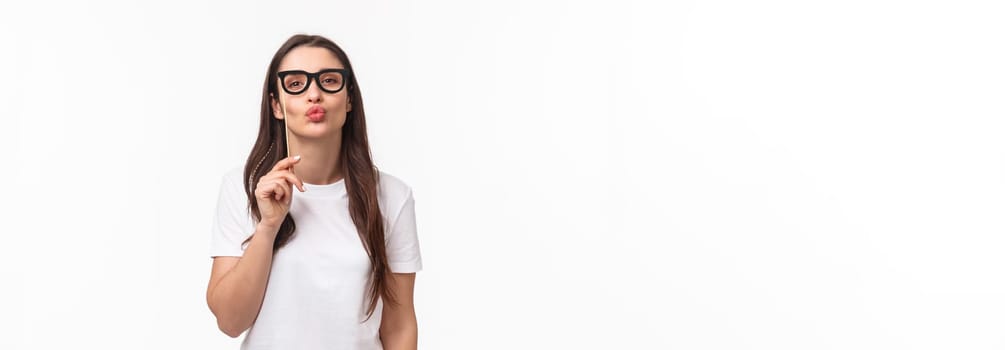 Entertainment, fun and holidays concept. Portrait of silly and cute young attractive girl wearing carnaval mask glasses, folding lips to give air kiss, pouting and standing white background.