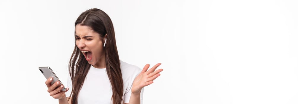 Close-up portrait of pissed-off aggressive and angry young brunette woman yelling at person while having phone call in wireless earphones, screaming at smartphone outraged.