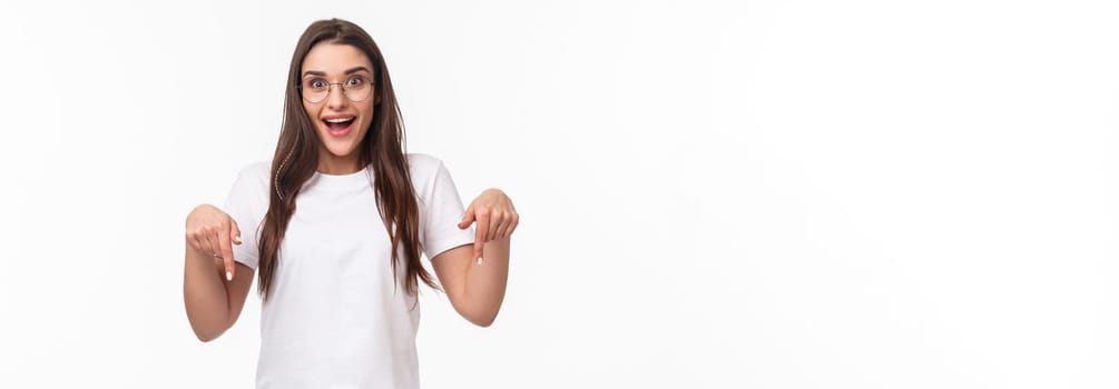 Waist-up portrait of excited young pretty woman seeing fantastic news, event promo or special spring discount, pointing fingers down and looking camera enthusiastic, stand white background.