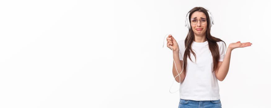 Technology, lifestyle and music concept. Portrait of awkward and frustrated young gloomy girl in headphones, holding wire and shrugging upset, earphones broke, want buy wireless earbuds.