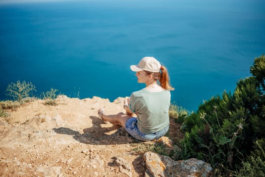 A woman sits on a rocky hillside overlooking the ocean. She is wearing a green shirt and white hat