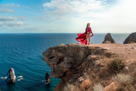 A woman in a red dress stands on a rocky cliff overlooking the ocean. The scene is serene and peaceful, with the woman's flowing dress adding a sense of grace and beauty to the landscape