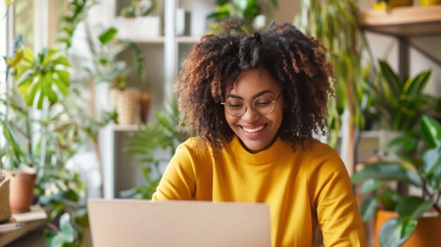 Happy modern young african woman sitting at desk and working at the laptop at home with green plants, black female freelancer or student