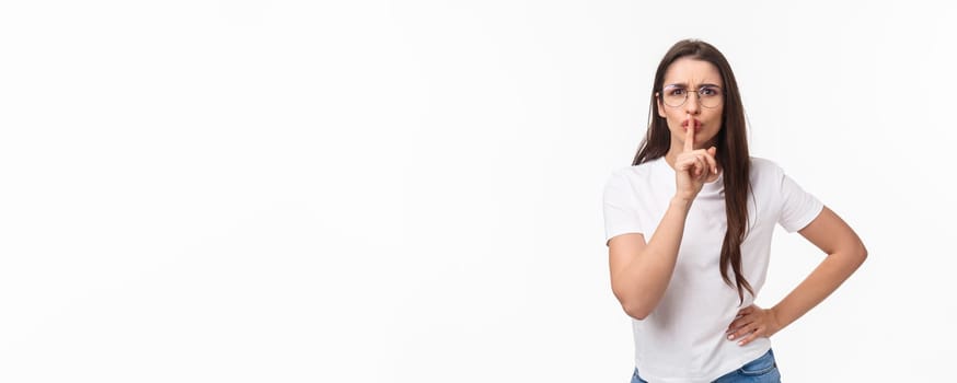 Waist-up portrait of angry and disappointed, serious-looking brunette woman shushing at camera with irritated face, telling keep quiet during lecure, hush with finger pressed to lips.