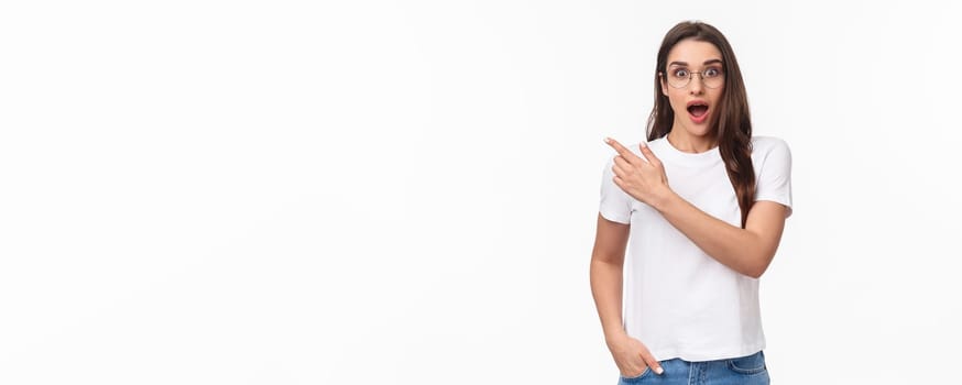 Waist-up portrait of enthusiastic, amazed and impressed young attractive girl in glasses talking about amazing new event or promo she saw online, pointing finger upper left corner, gasping astonished.