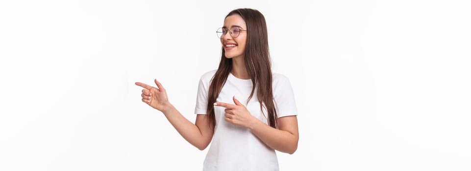 Waist-up portrait of cheerful young female student, programmer, pointing fingers and looking at right copy space, smiling and laughing as seeing something entertaining and funny, white background.