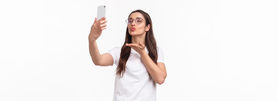 Communication, technology and lifestyle concept. Lovely attractive woman in glasses, sending air kiss to her followers online live stream with app, taking photo with filters, hold mobile phone.