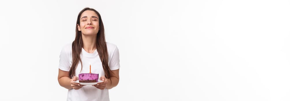 Entertainment, fun and holidays concept. Portrait of romantic beautiful young b-day girl celebrating throwing party, holding piece of birthday cake with candle, making wish with smile and closed eyes.