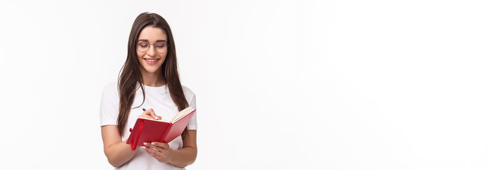 Portrait of smiling pretty caucasian girl writing in her diary, prepare homework for english courses, smiling, have inspiration to write plans for next week in planner, white background.