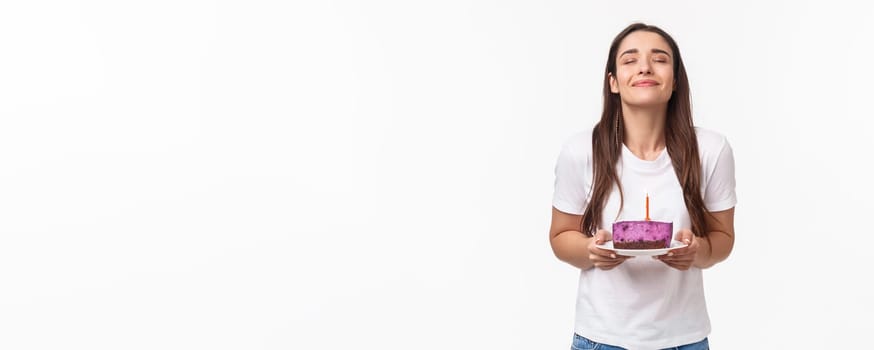 Entertainment, fun and holidays concept. Portrait of romantic beautiful young b-day girl celebrating throwing party, holding piece of birthday cake with candle, making wish with smile and closed eyes.