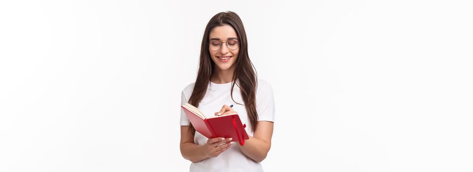 Portrait of smiling pretty caucasian girl writing in her diary, prepare homework for english courses, smiling, have inspiration to write plans for next week in planner, white background.