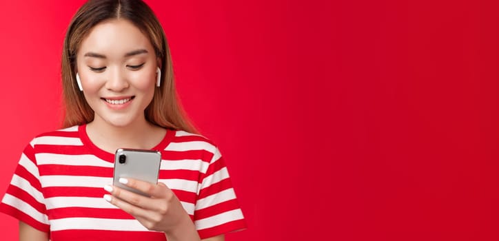 Carefree modern asian blond girl picking song wear wireless earphones, listen music using online song platform app, hold smartphone look pleased smiling phone screen, stand red background.