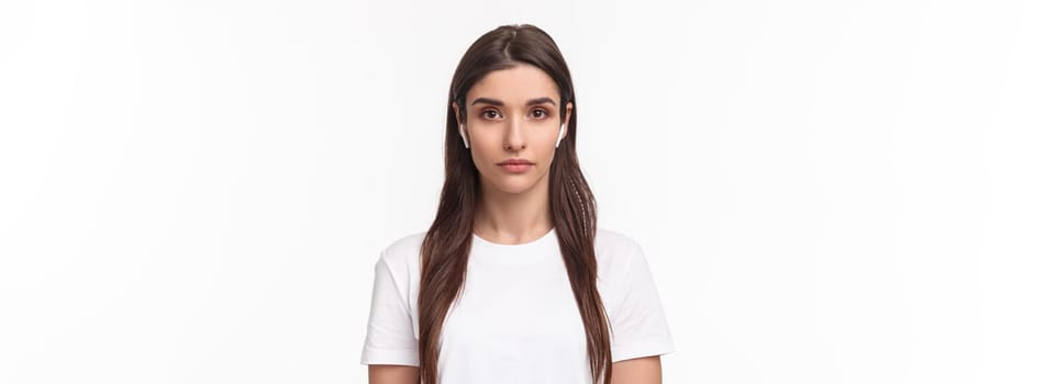 Close-up portrait of serious-looking attractive caucasian woman in wireless earphones, looking at camera with relaxed no emotions face, standing white background. Copy space