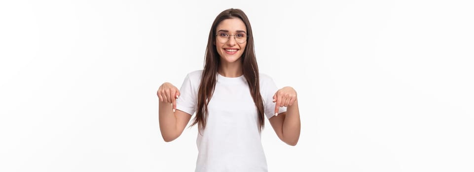 Click here. Portrait of brunette gorgeous young woman in glasses, pointing fingers down to show advertisement, smiling pleased, suggest visit link, recommend download or subscribe, white background.