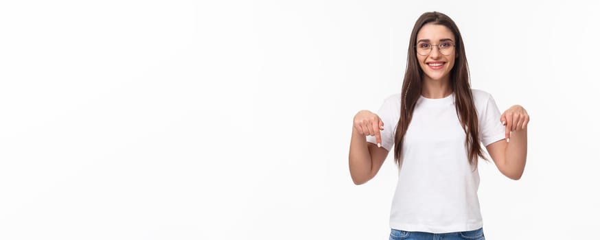 Click here. Portrait of brunette gorgeous young woman in glasses, pointing fingers down to show advertisement, smiling pleased, suggest visit link, recommend download or subscribe, white background.