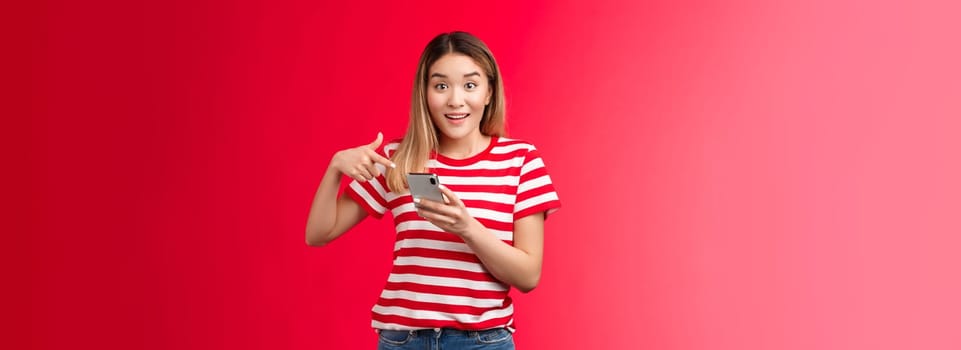 Girl telling you about awesome promo online shop, smiling impressed hold smartphone pointing finger phone screen, discuss new friend post surprised pleased, stand red background amazed.