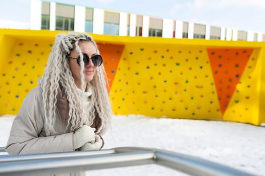 A woman with long white hair is wearing sunglasses. She is standing outdoors and looking ahead with a serious expression on her face. The sun is shining, reflecting off her glasses.