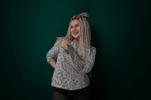 A woman with dreadlocks stands confidently in front of a bright green wall, looking directly at the camera. Her hair is styled in long, thick dreadlocks, and she wears a casual outfit. The background is a solid green color, providing a striking contrast to the womans appearance.