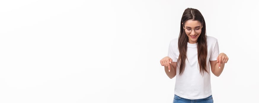 Waist-up portrait of enthusiastic, smiling happy brunette female student in glasses and t-shirt, looking pointing down, bottom advertisement, glancing pleased, standing white background.