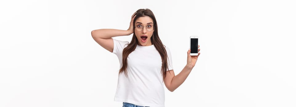 Communication, technology and lifestyle concept. Waist-up portrait of astonished surprised and excited young woman in glasses, gasping and touch head shocked as showing mobile phone awesome app.