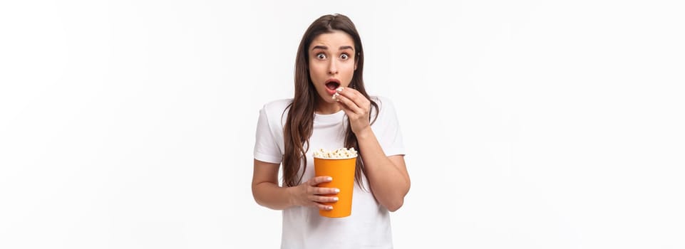 Entertainment, fun and holidays concept. Portrait of astonished and excited impressed young girl watching awesome movie, look astonished eating popcorn, pop eyes at tv screen, white background.