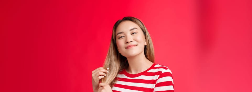 Close-up satisfied cute carefree asian blond girl feeling delighted and pleased after haircare procedure touch healthy hair tilt head smiling broadly like new hairdresser, stand red background.