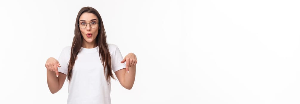 Portrait of amused and curious brunette female student inviting everyone check out very interesting event, follow this page or link, visit store, pointing fingers down, bottom advertisement.